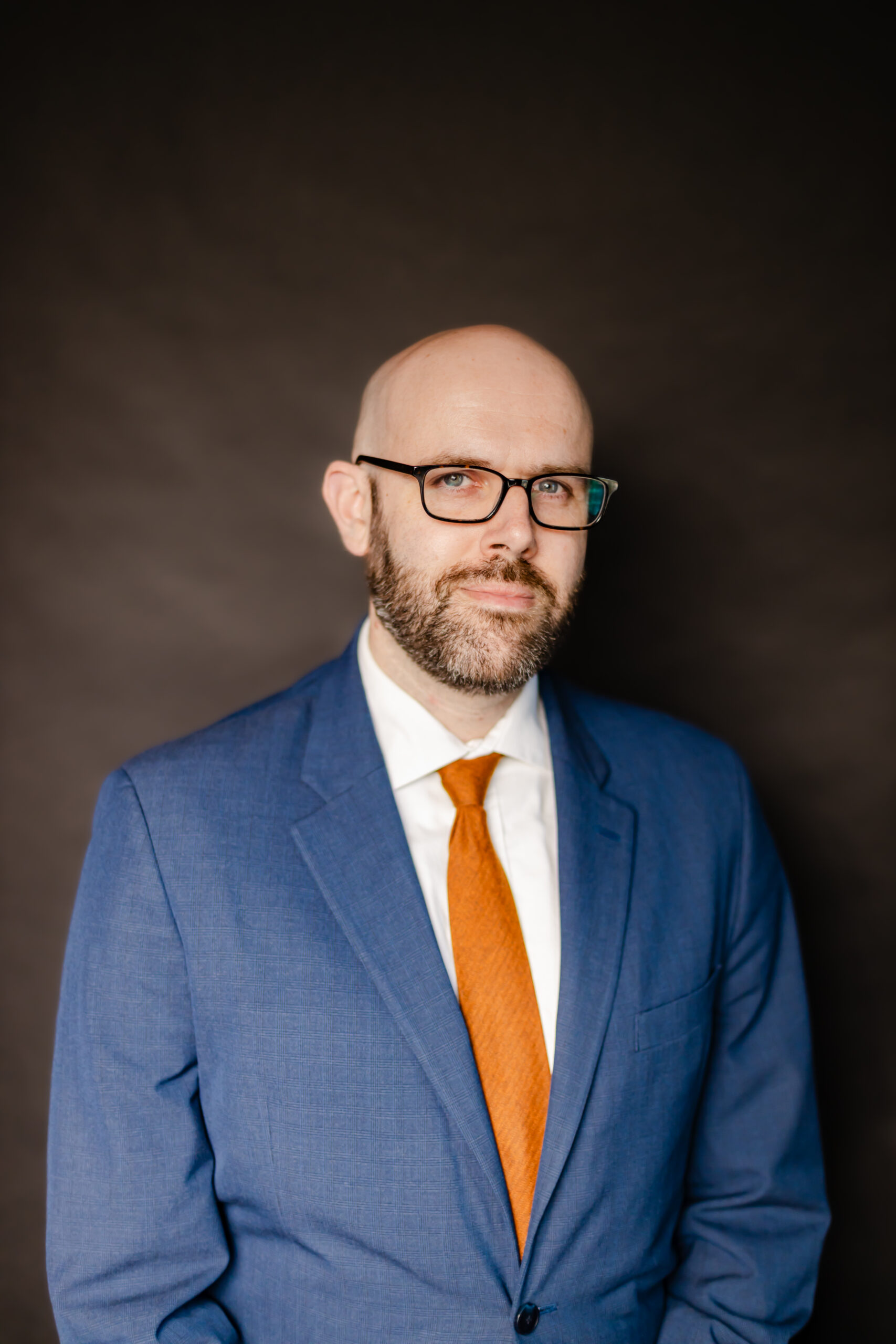 Professional portrait of a male defense attorney wearing a blue suit and orange tie against a dark studio background.